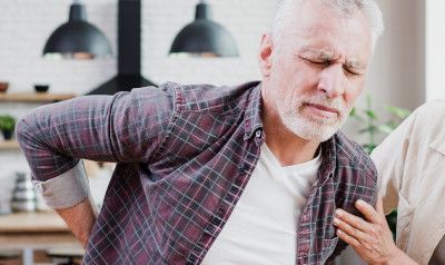 Man in pain, clutching his lower back, indoors. Another person's hand on his arm.