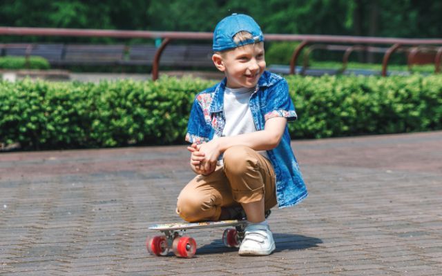 Young boy kneels on skateboard, smiling. Wearing blue cap, shirt, brown pants, white shoes, outdoors.