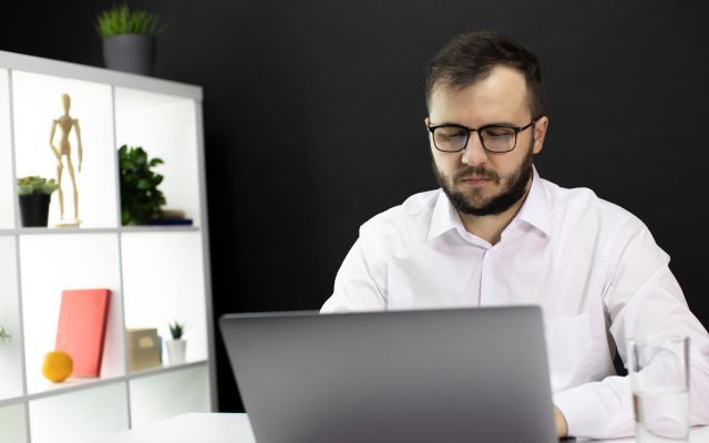 Man with glasses working on a laptop in a modern office, looking intently at the screen.