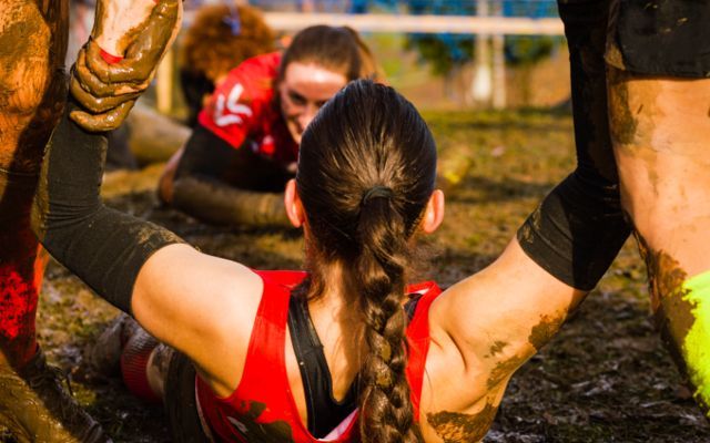 Woman in red shirt crawls through mud during an obstacle course, being aided by others.