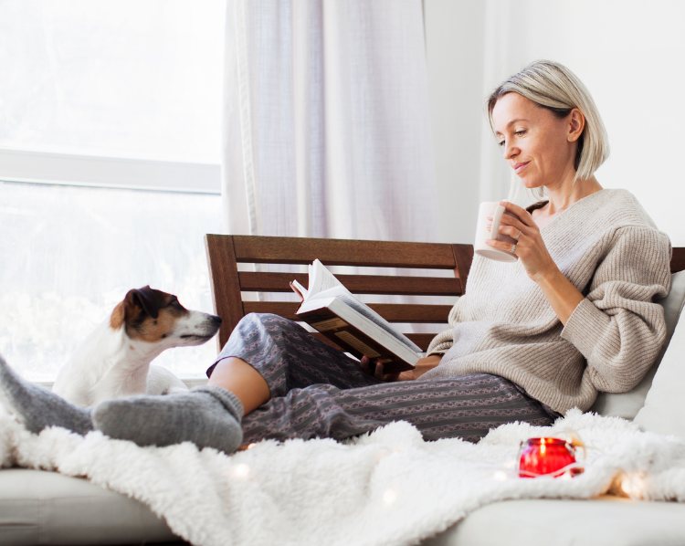 Woman reading, drinking coffee on couch with dog. Cozy setting, neutral tones.