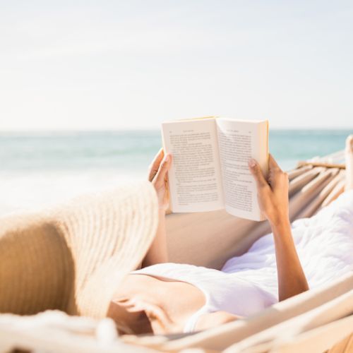 Person lying in a hammock reading a book on the beach, with the ocean in the background.