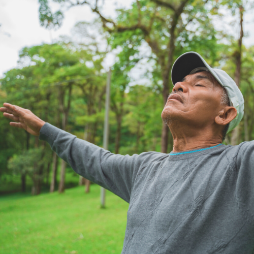 Man with arms outstretched, eyes closed, in a green field with trees; looks relaxed, hopeful.