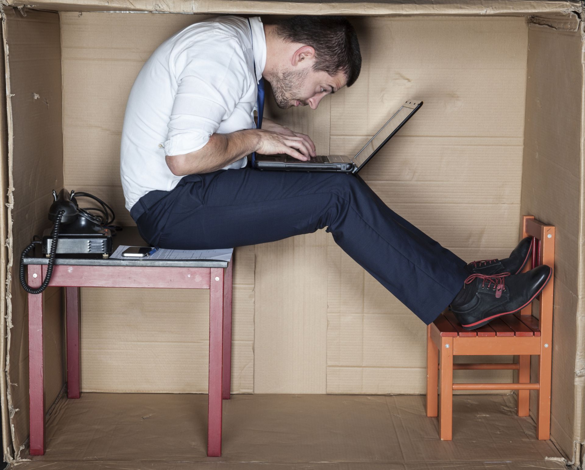 Man hunched over laptop in a small box, legs stretched. Desk and chair are visible.