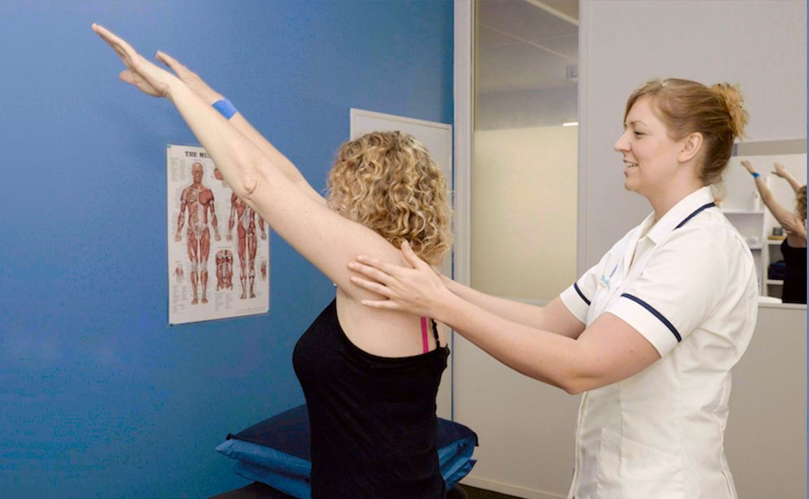 Woman has arm raised while a nurse supports it, indoors with blue wall.