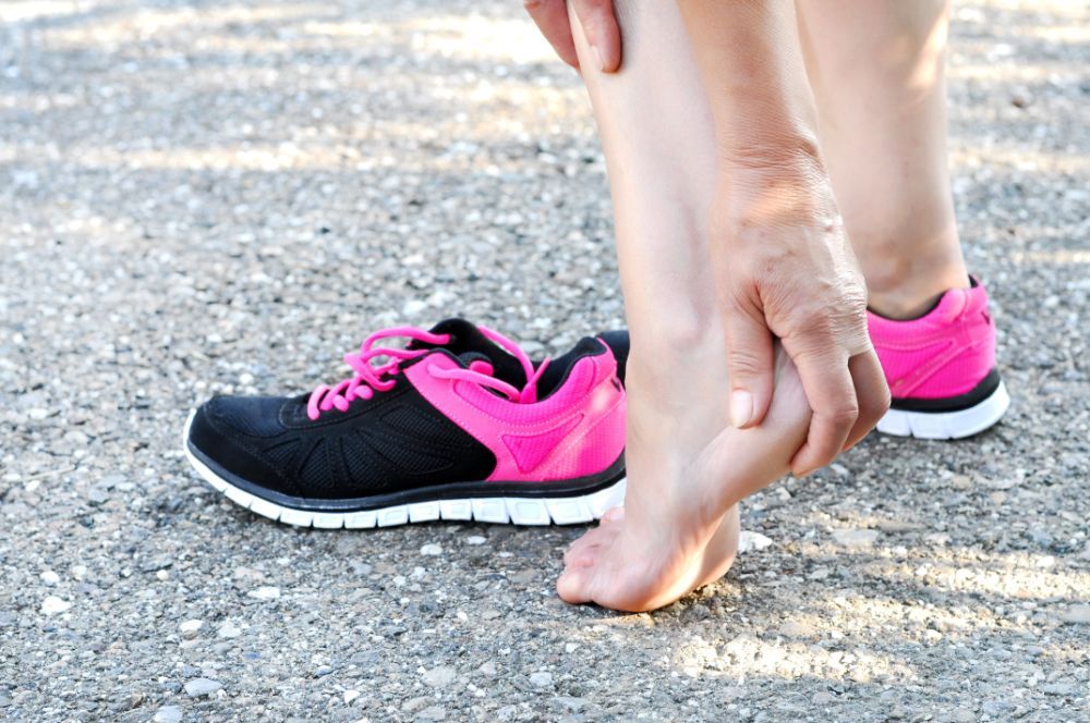 Person holding their ankle in pain next to a pink and black running shoe on a gravel surface.