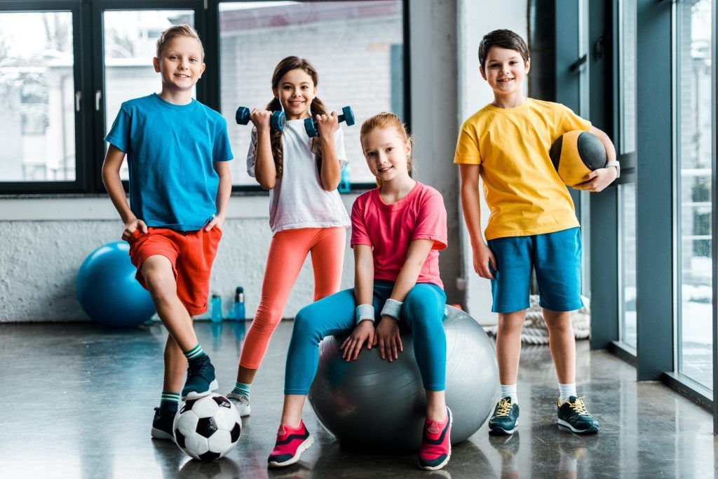 Group of children in a gym; one holds dumbbells, one sits on a ball, another holds a ball, and one has a soccer ball.