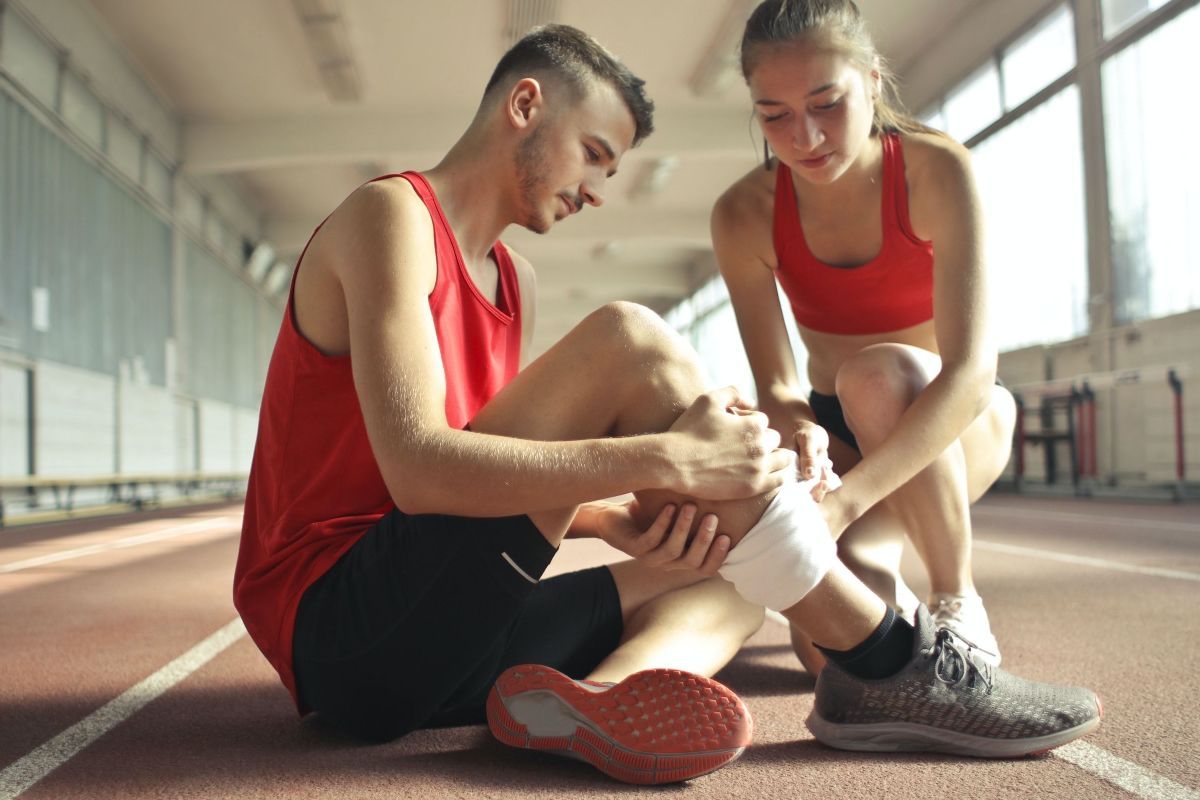 Man with bandaged ankle is assisted by woman on a running track. Both are wearing red athletic attire.