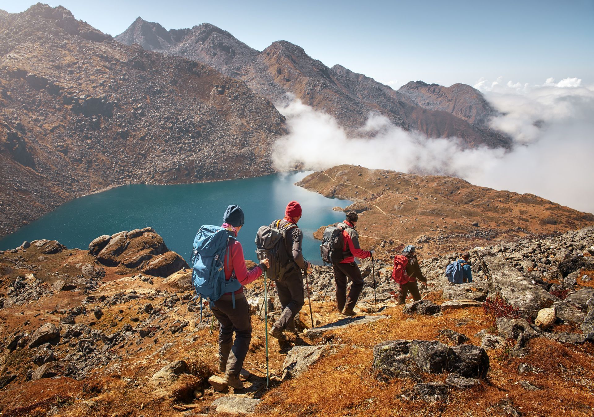 Hikers trek on a rocky mountain path, overlooking a lake and mountain range.