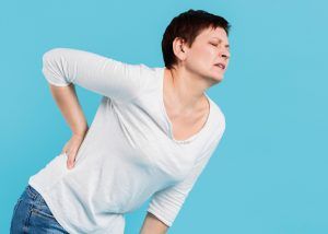Woman in white shirt, clutching back, showing pain, against a blue background.