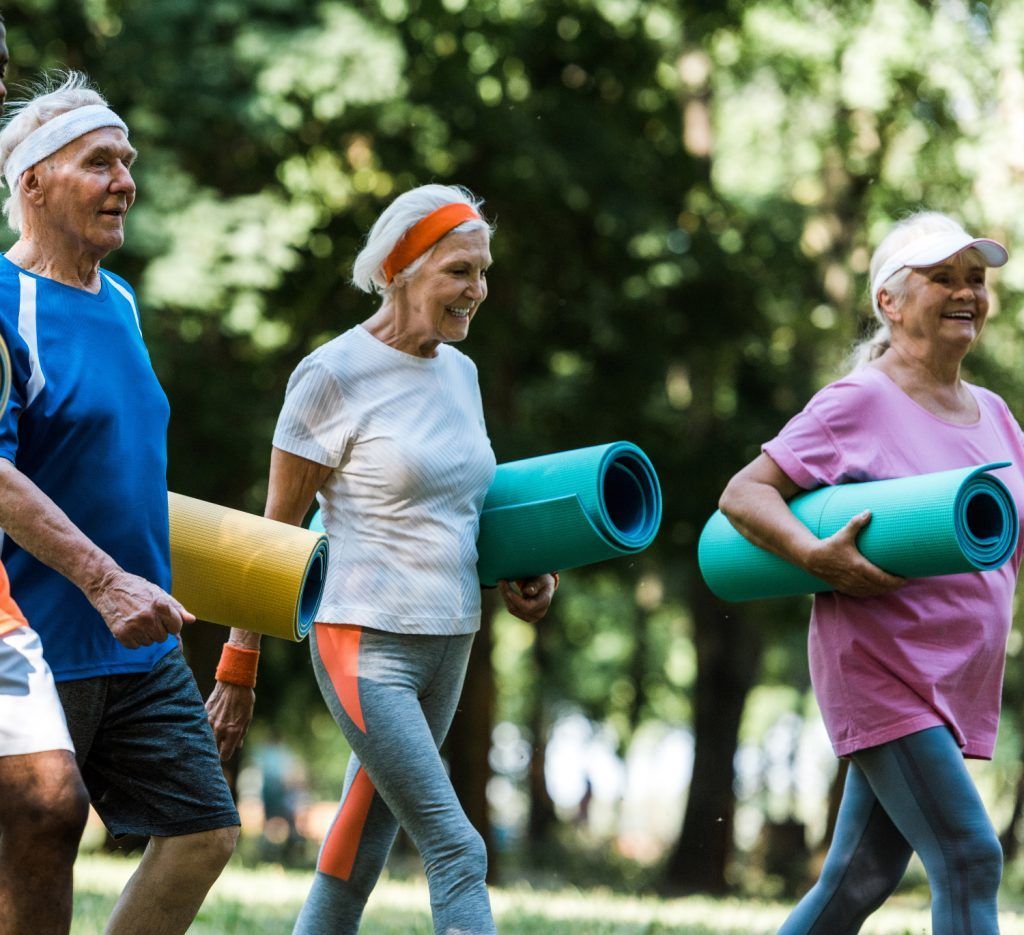 Three older adults walking outdoors, each carrying a rolled-up yoga mat.
