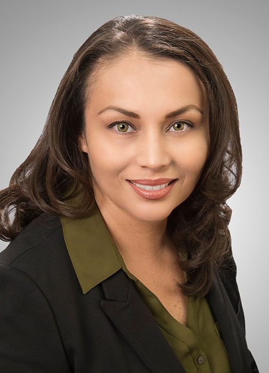Woman with brown hair, smiling, wearing a black blazer and olive green shirt.