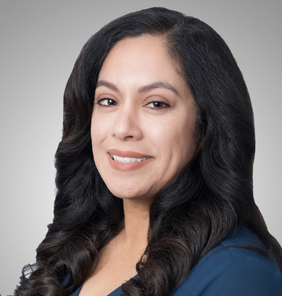 Woman with dark hair smiles at the camera, wearing a blue top against a gray backdrop.