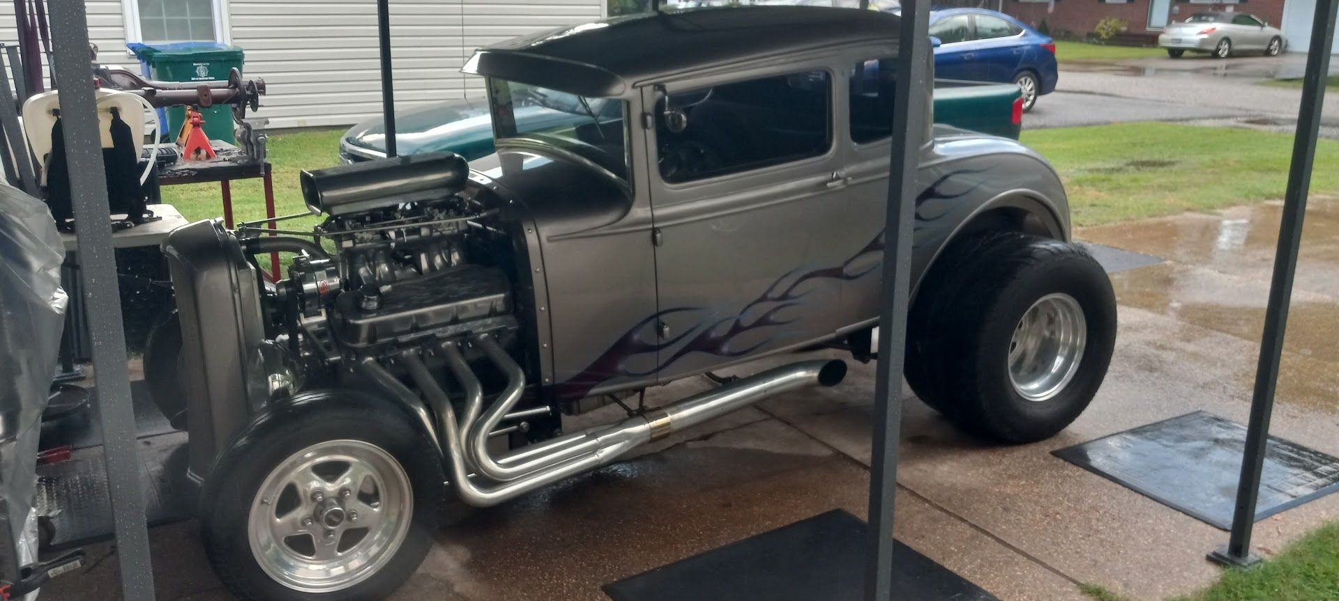 A silver classic hot rod with large rear tires, an exposed engine, and black flame graphics parked under a shelter | Sprinkle and Williams II, Inc.