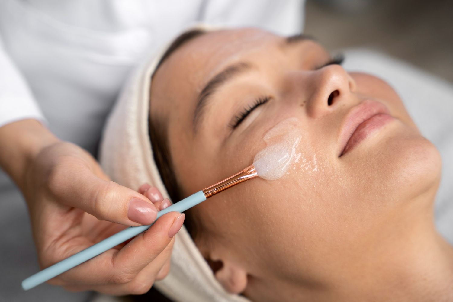 Woman receiving facial treatment; a person applies product with a brush.