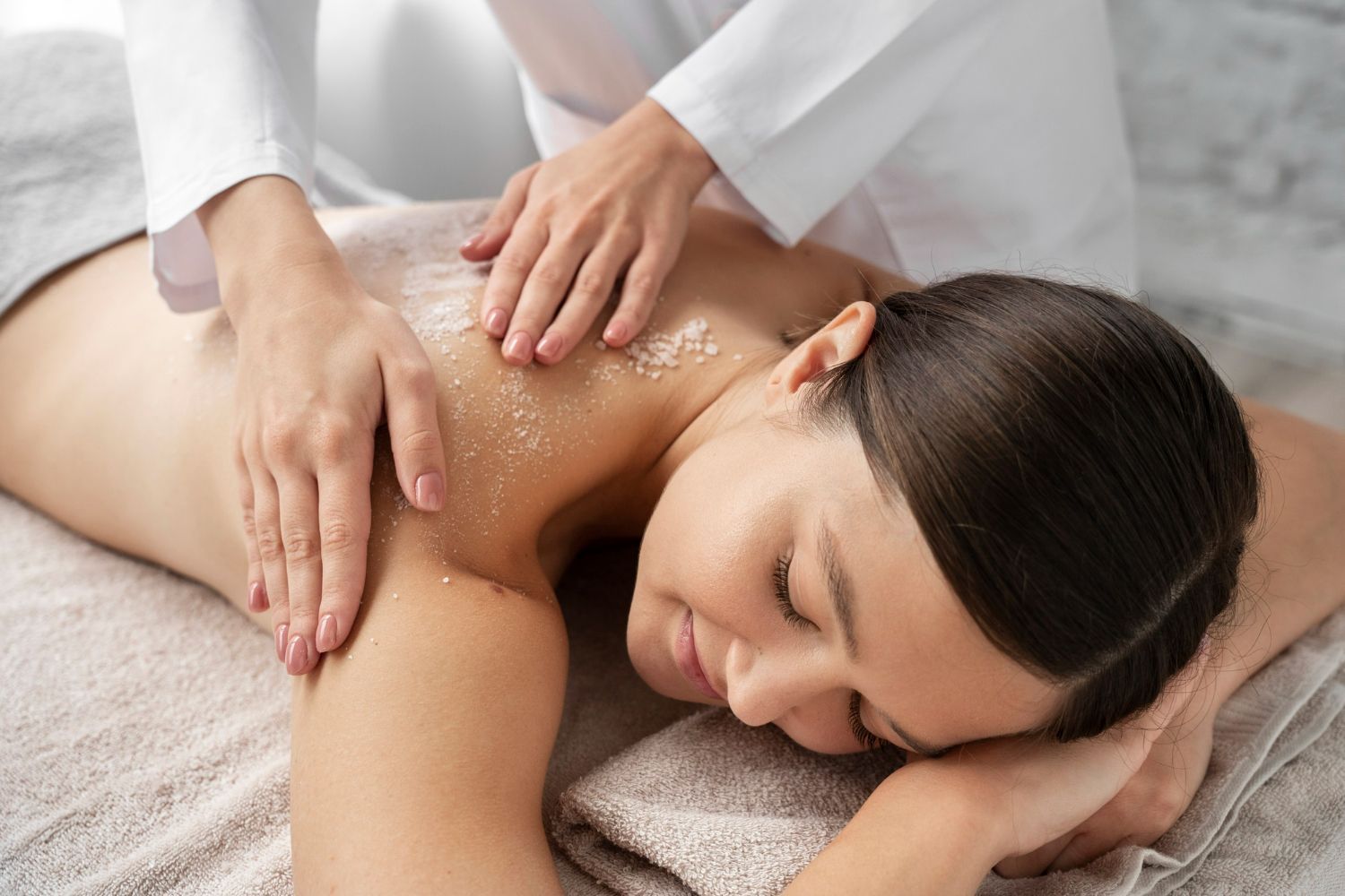 Person receiving back scrub, spa setting. Hands on back, light skin, closed eyes, smiling.