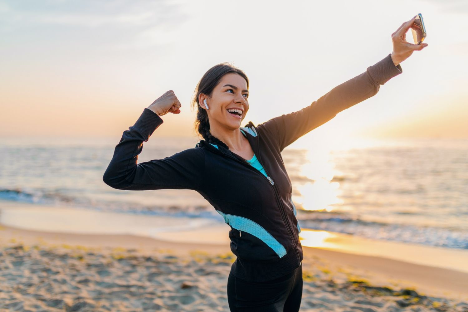 Woman takes selfie on beach at sunrise, flexing bicep, wearing activewear.