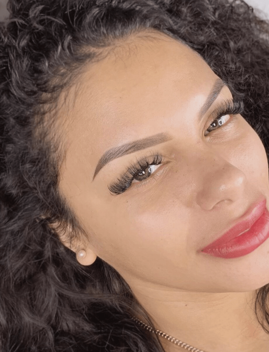 Woman with dark curly hair and bold makeup smiles, showcasing eyebrow and lip enhancements.