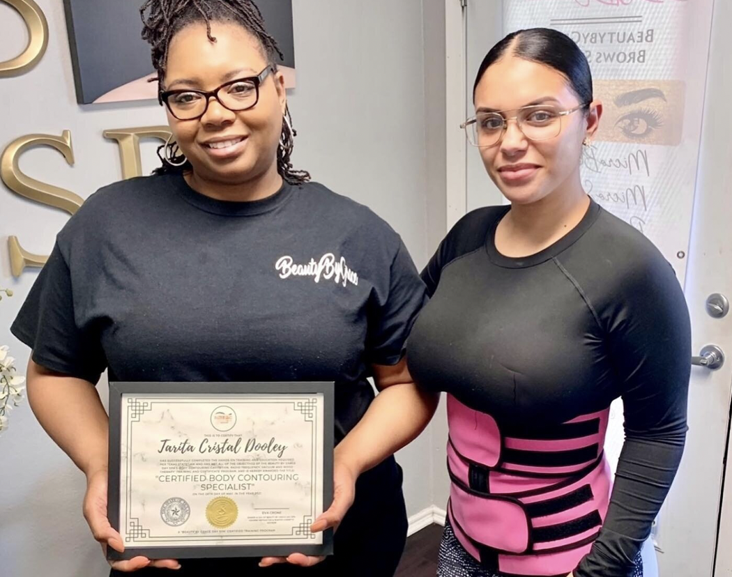 Two women hold a certificate. One wears a black t-shirt, the other a pink waist trainer. They are in a studio.