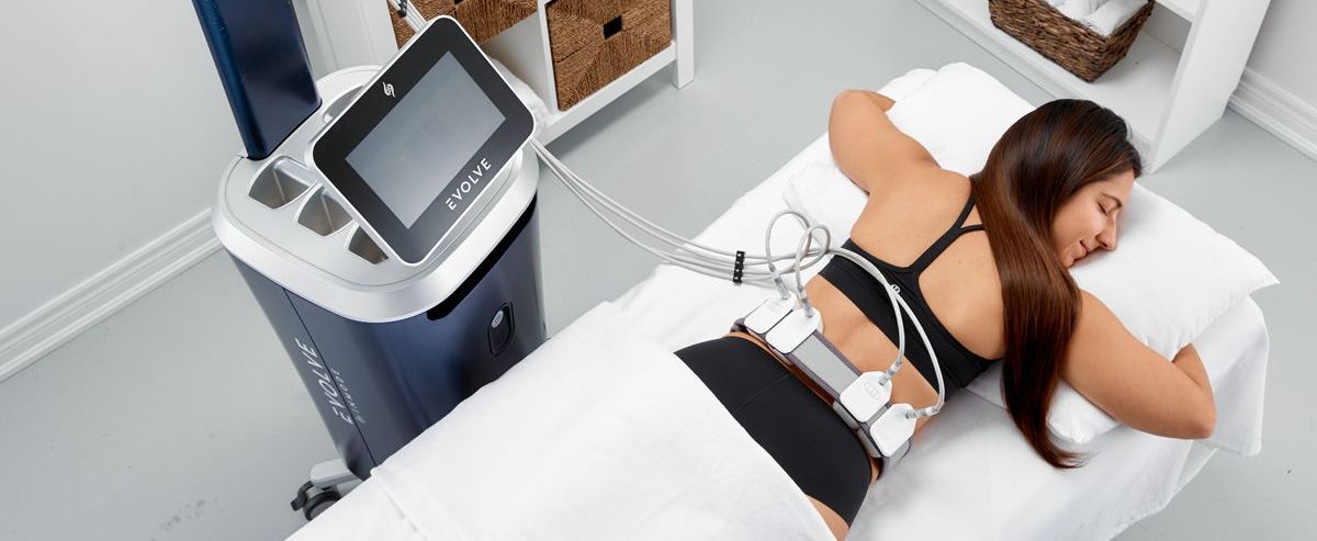 A woman lies face down on a spa bed, undergoing a body contouring treatment.  She has applicators on her back and sides, and is next to a blue medical device.
