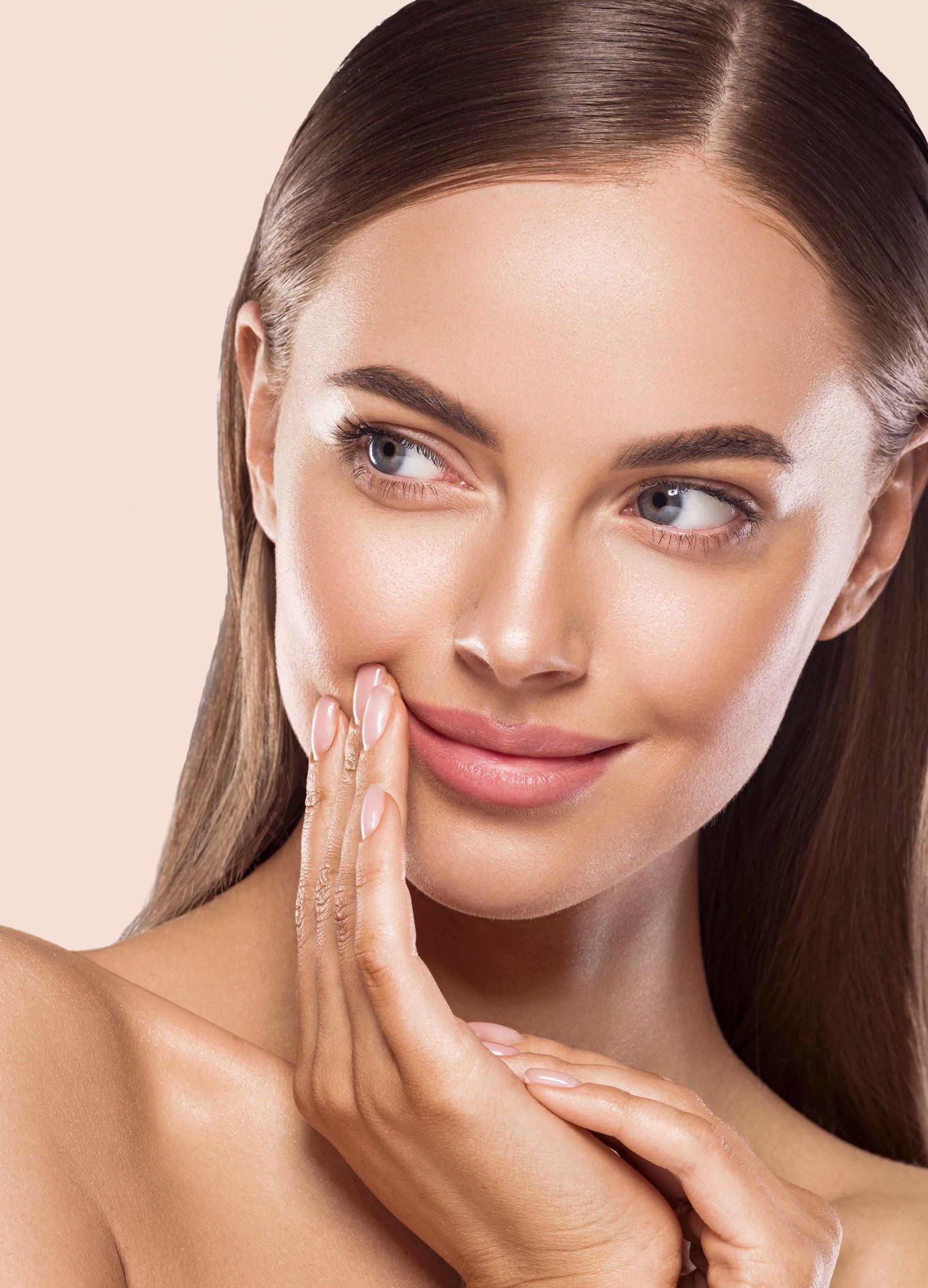 Woman with light skin touching her cheek with fingers, smiling, against a tan background.
