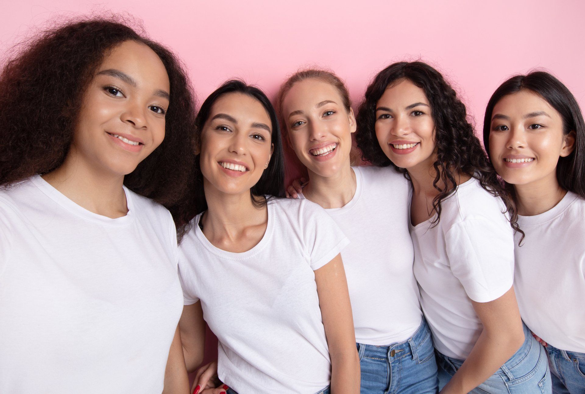 Five people in white shirts and jeans smiling, against a pink background.