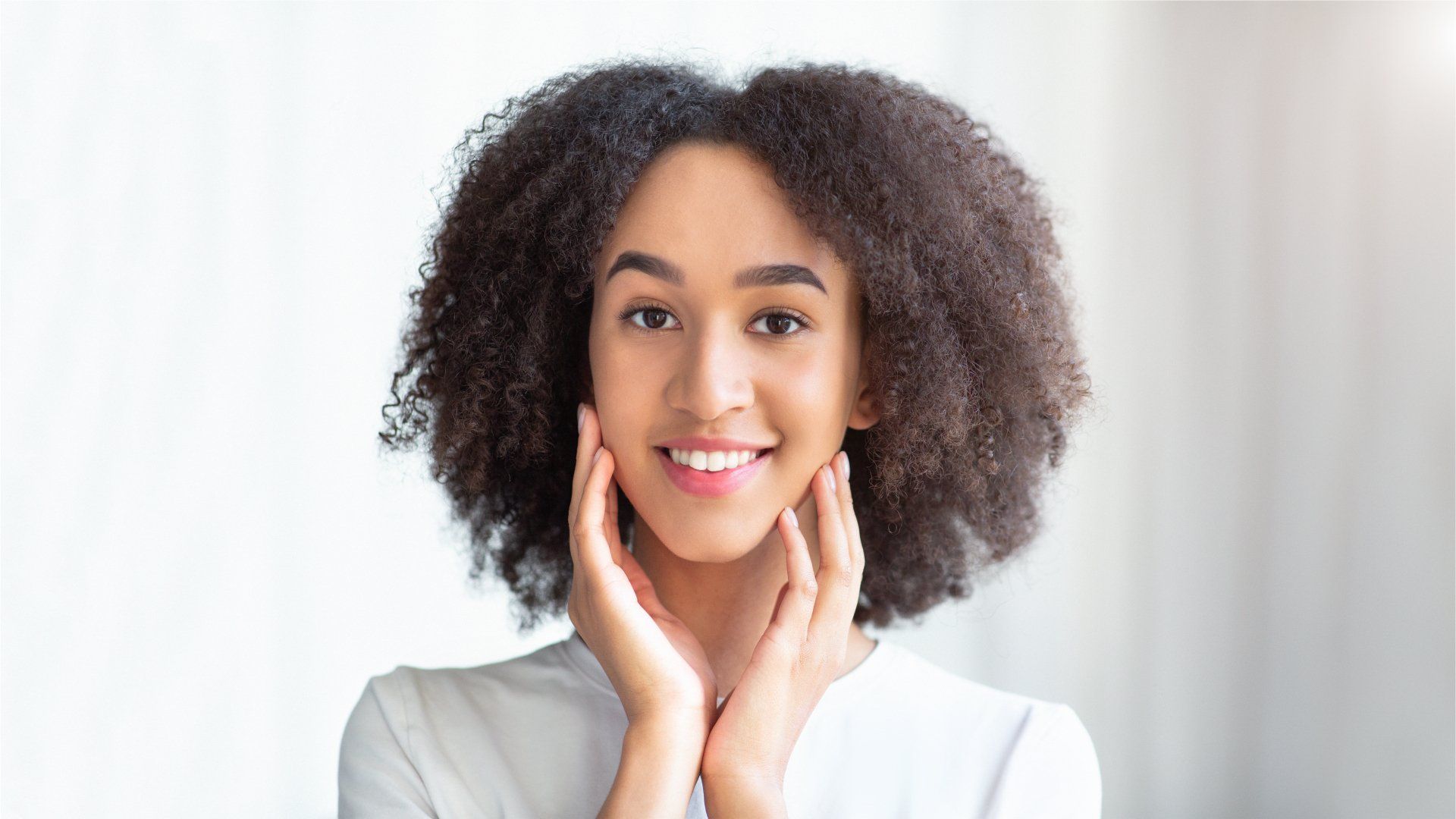 Woman with curly dark hair smiles, touching her cheeks, against a white background.