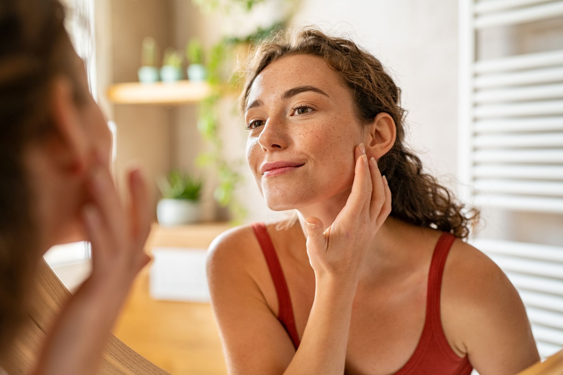 Woman in a red top looks at her face in a bathroom mirror, touching her cheek with a smiling expression.