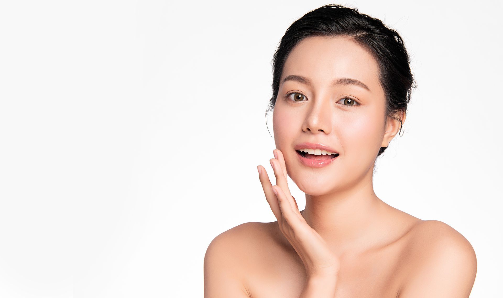 Woman with fair skin, smiling, touching her cheek. Against a white background.