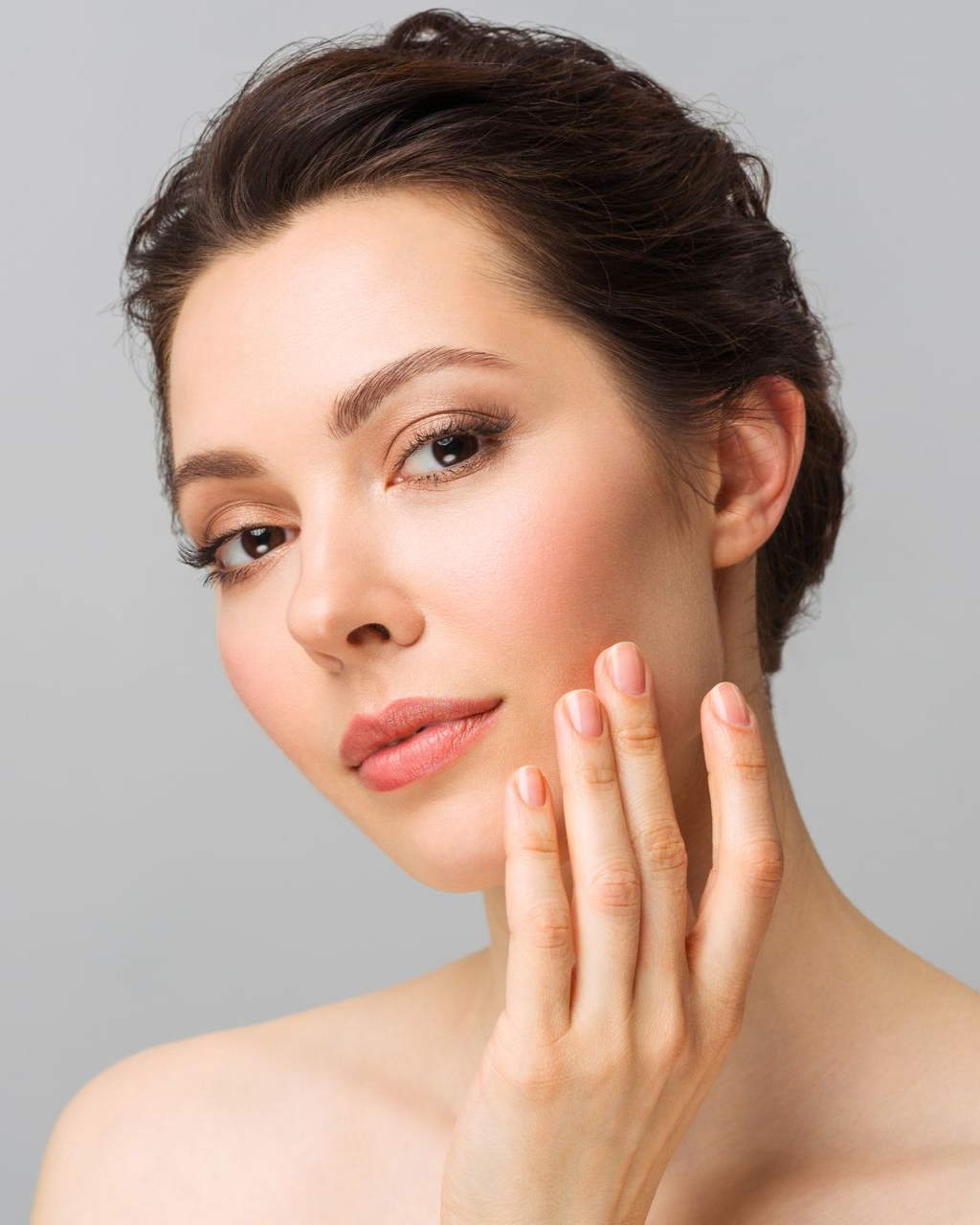 Woman touching her face, looking at camera with a soft expression. Light makeup, fair skin, gray background.