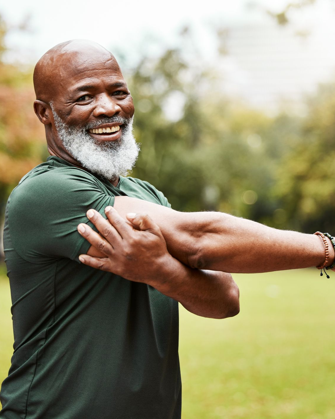 Man stretching arm outdoors, smiling. Green shirt, blurred background.