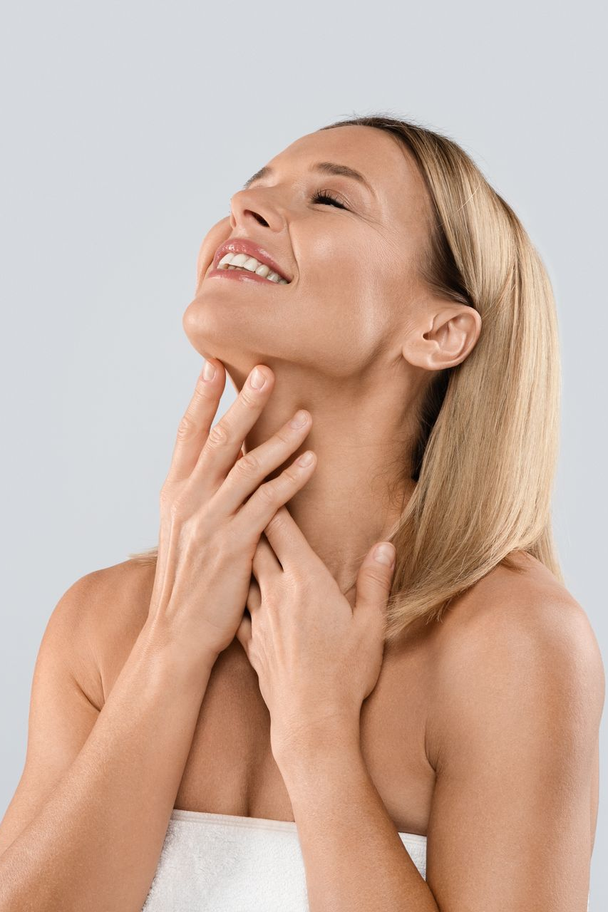 Woman smiles, touches neck with hands, eyes closed, against a light background.