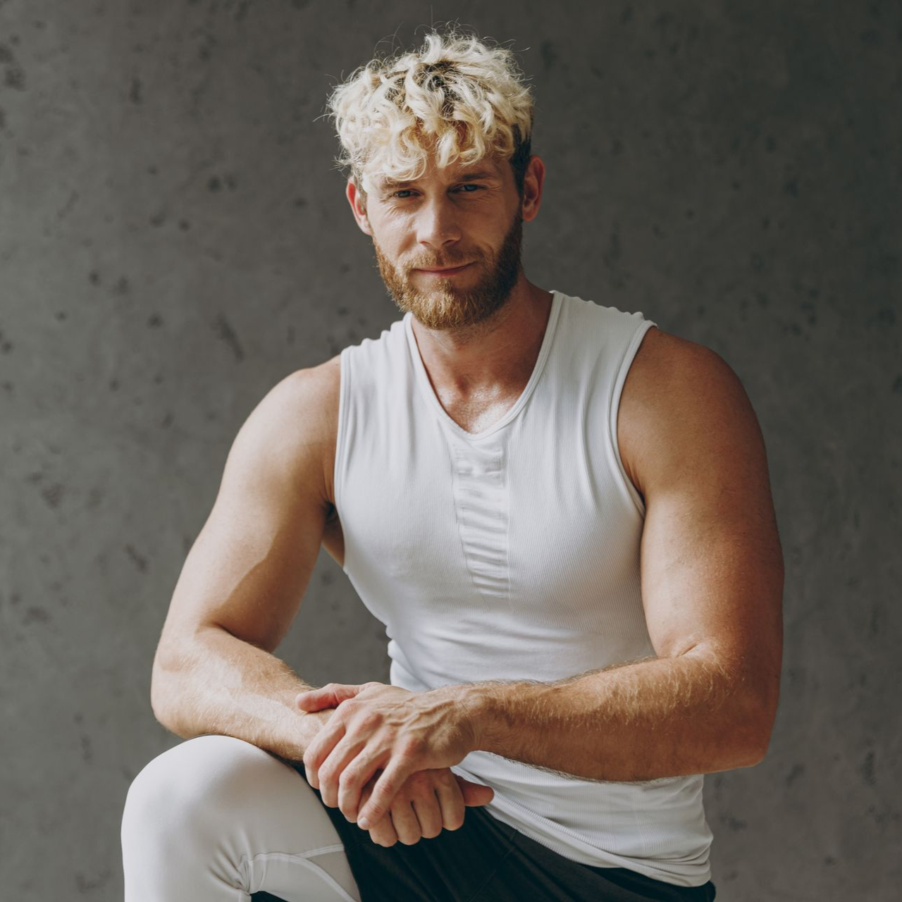 Man with blonde curly hair and beard, wearing a white tank top and kneeling, looking at the camera.