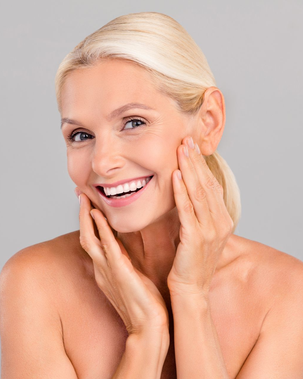 Blonde woman smiling, touching her cheeks with hands, neutral background.