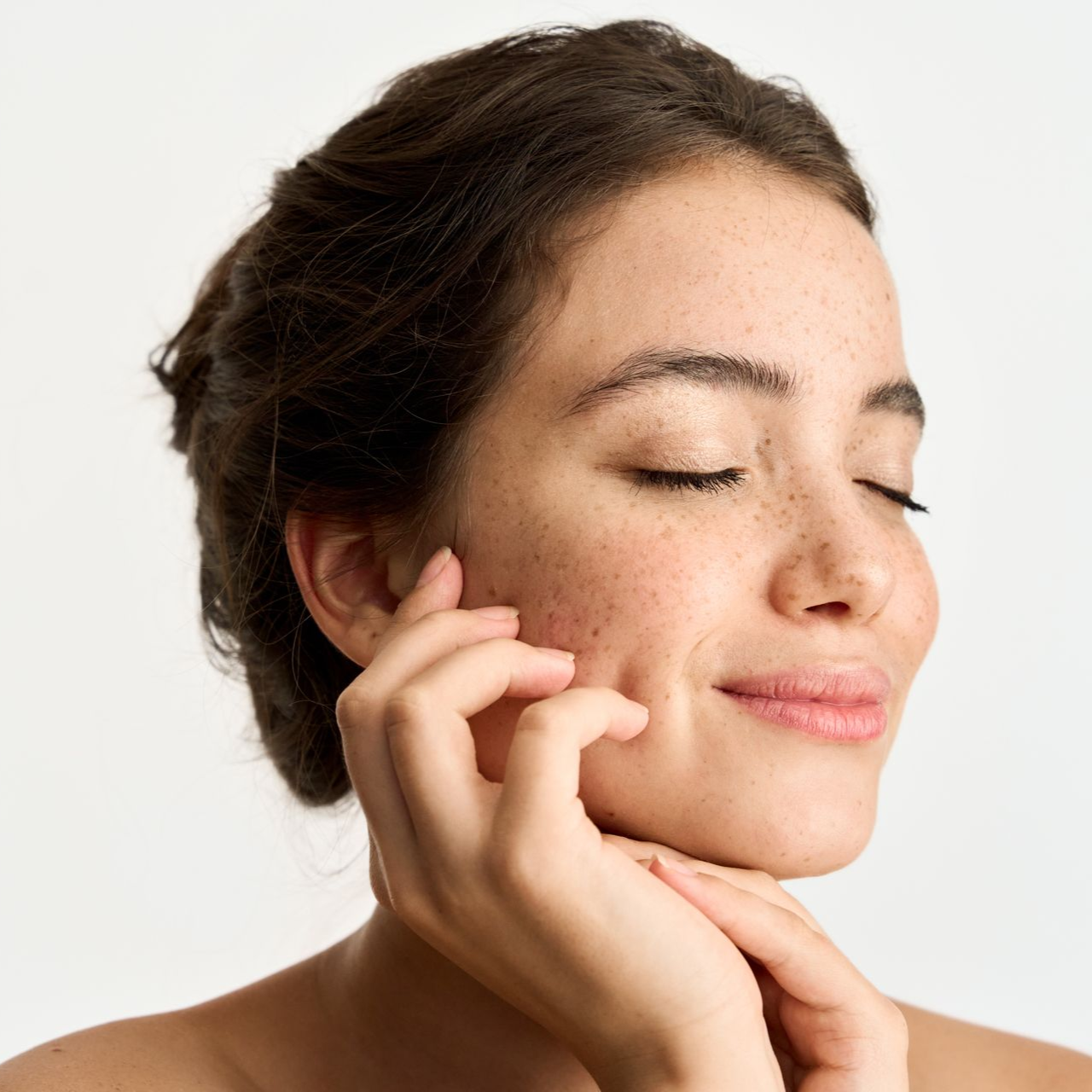 Woman with closed eyes, smiling, touches her face with both hands, small freckles visible.