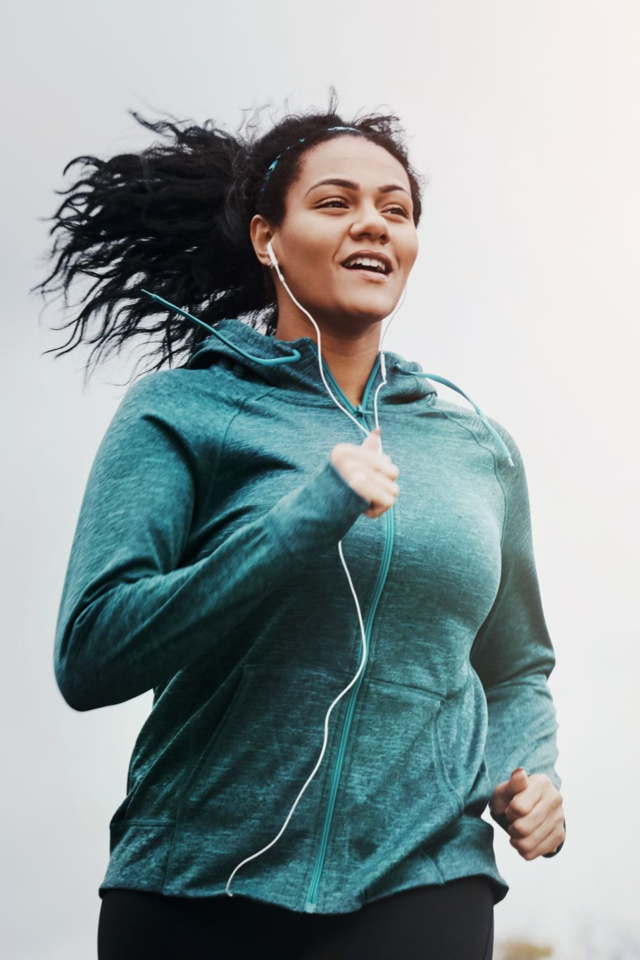 Woman jogging outdoors, wearing teal hoodie, earbuds, and smiling.