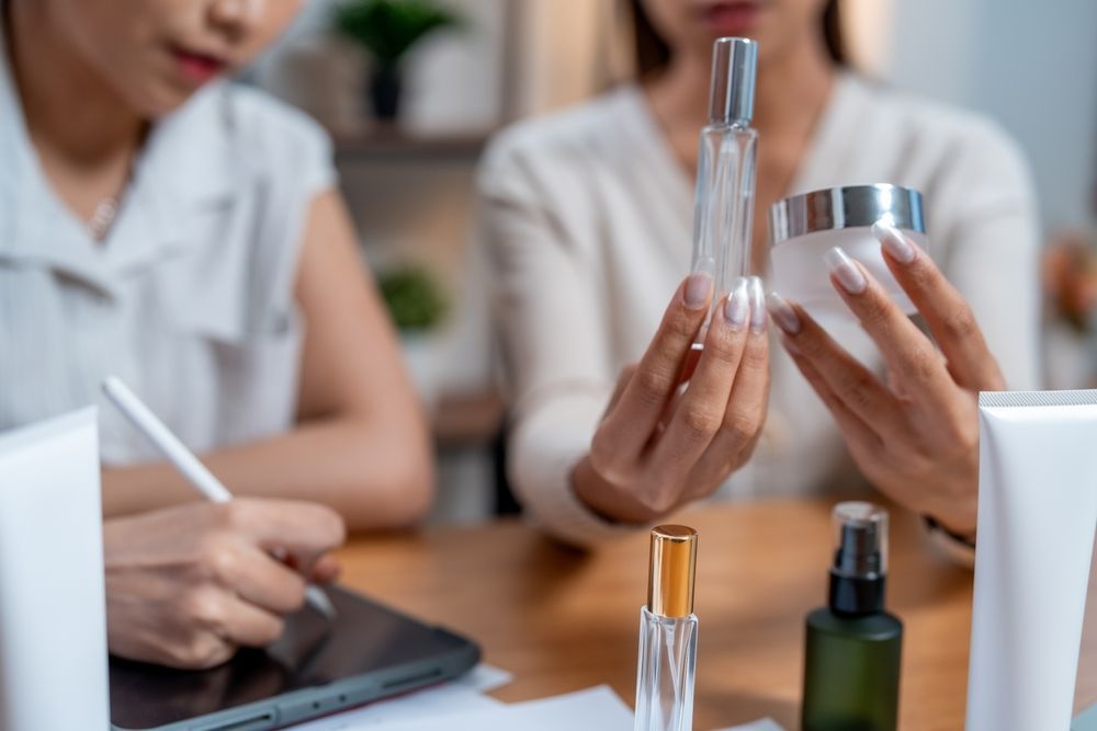 Two people examining cosmetic product bottles at a table; one person is writing on a tablet.