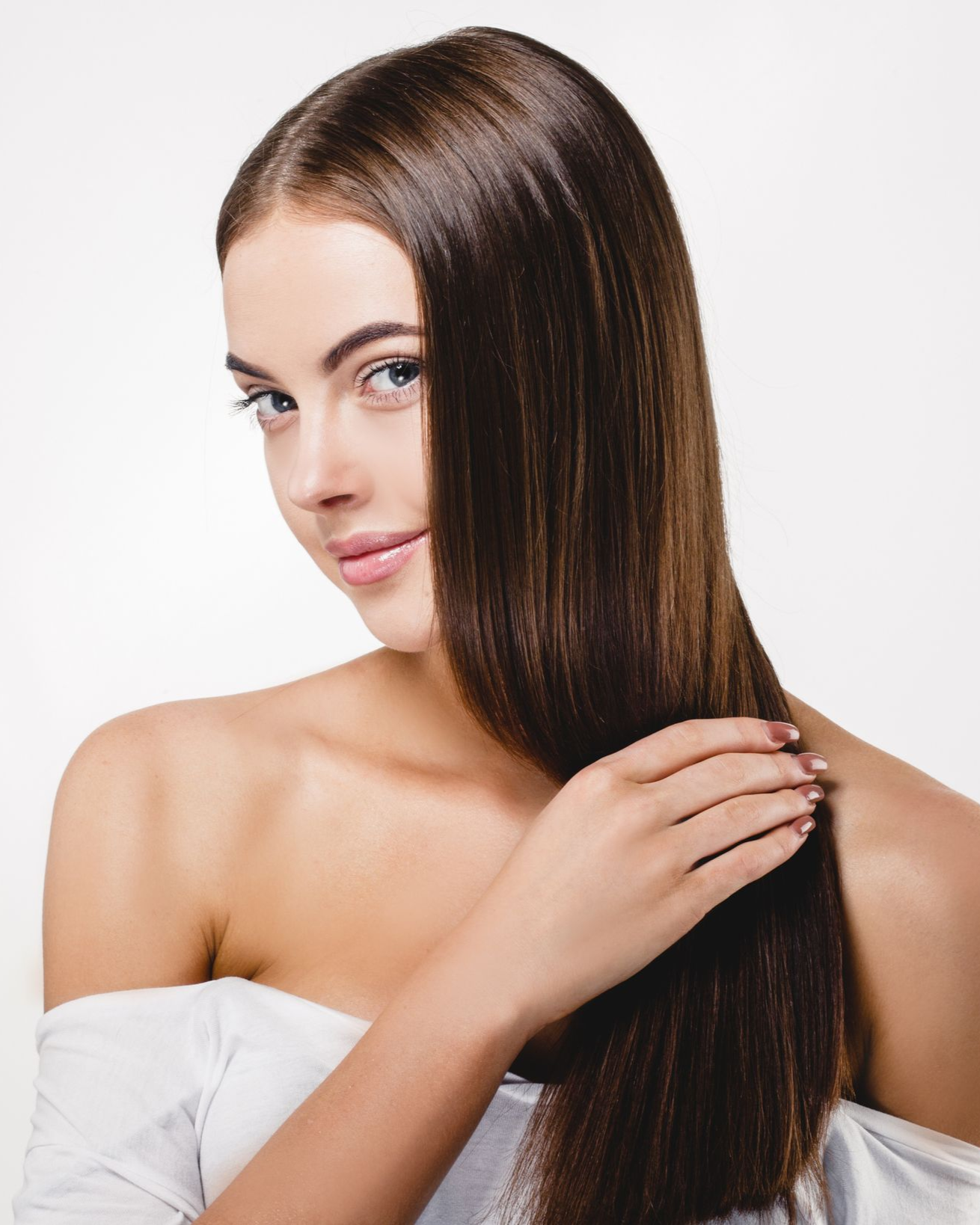 Woman with long, shiny, brown hair, touching it with her hand. She smiles, wearing a white top, against a white background.