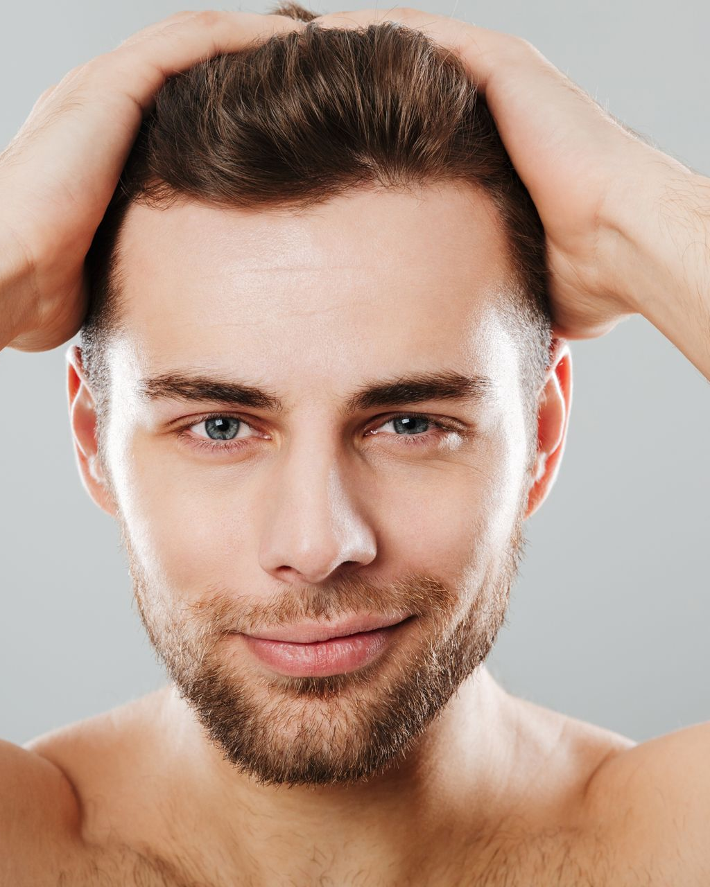 Man with hands on head, smiling. Brown hair and beard, blue eyes, neutral background.