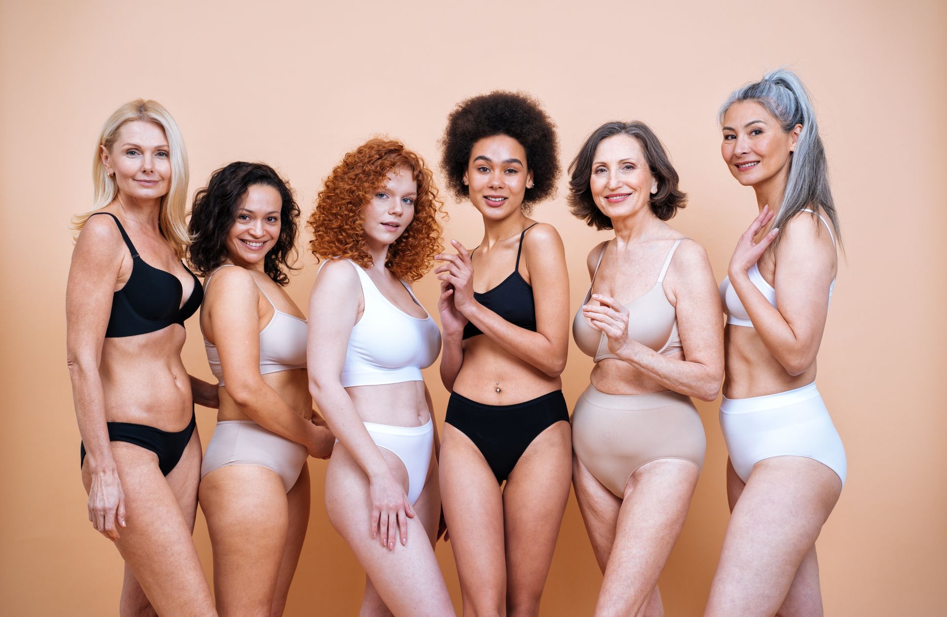Six diverse people in underwear stand against a neutral background, smiling and looking at the camera.