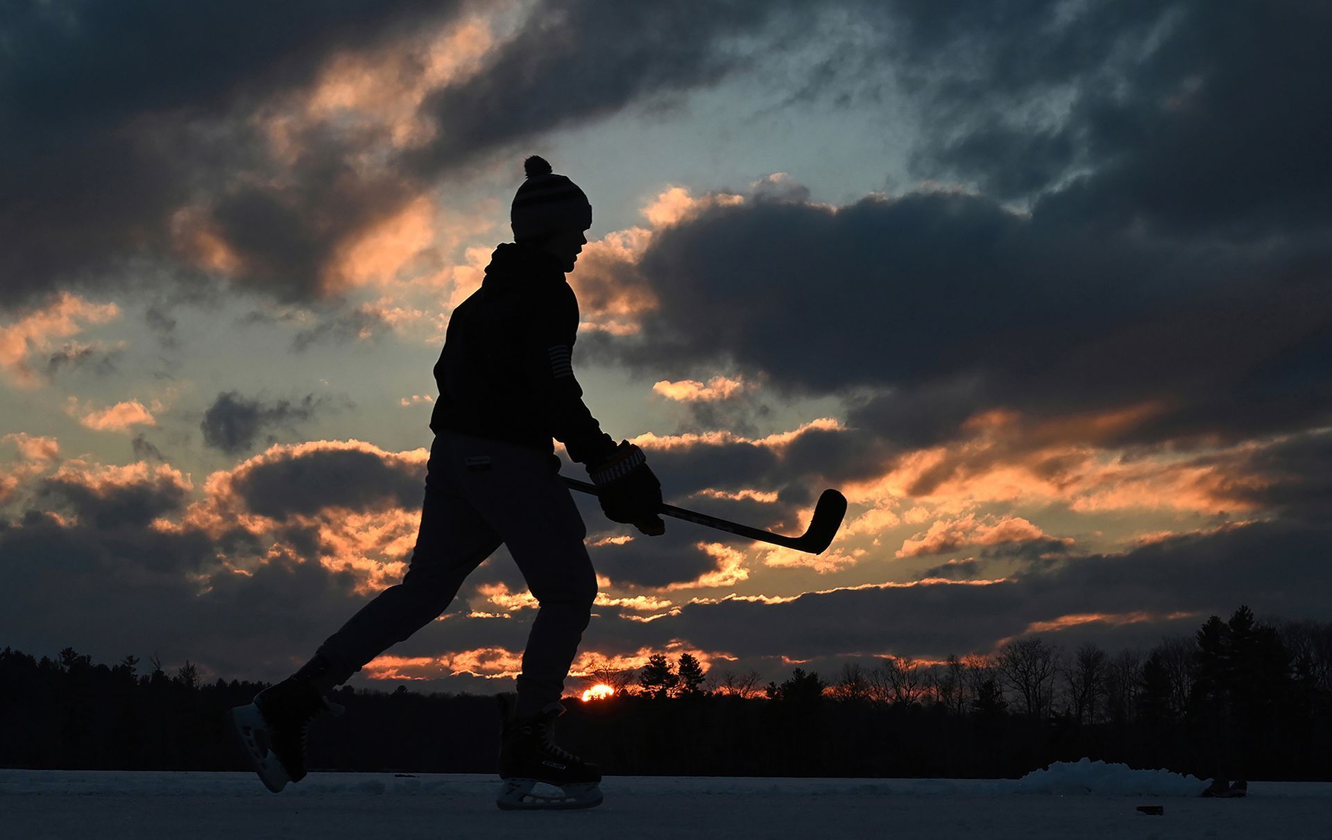 Jon Bowman, 15, is silhouetted by the setting sun as he waits for a pass from a friend while hitting a hockey puck around frozen Mill Pond in Westborough, Massachusetts, Jan. 25, 2022.