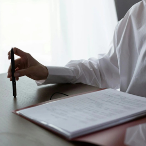 Person in white shirt holding a pen over an open notebook at a desk near a bright window