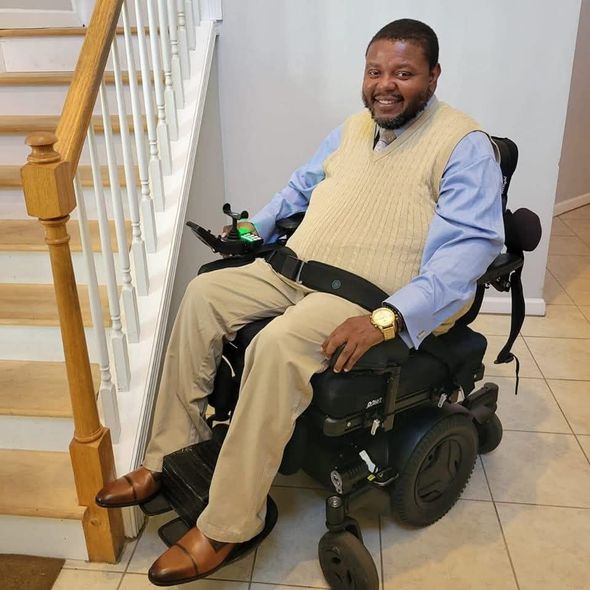 A smiling person in a light blue shirt and beige sweater vest sitting in a power wheelchair next to a wooden staircase.