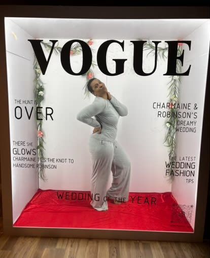 Woman posing in a Vogue-themed photo booth. She's in a gray jumpsuit on a red carpet, with 