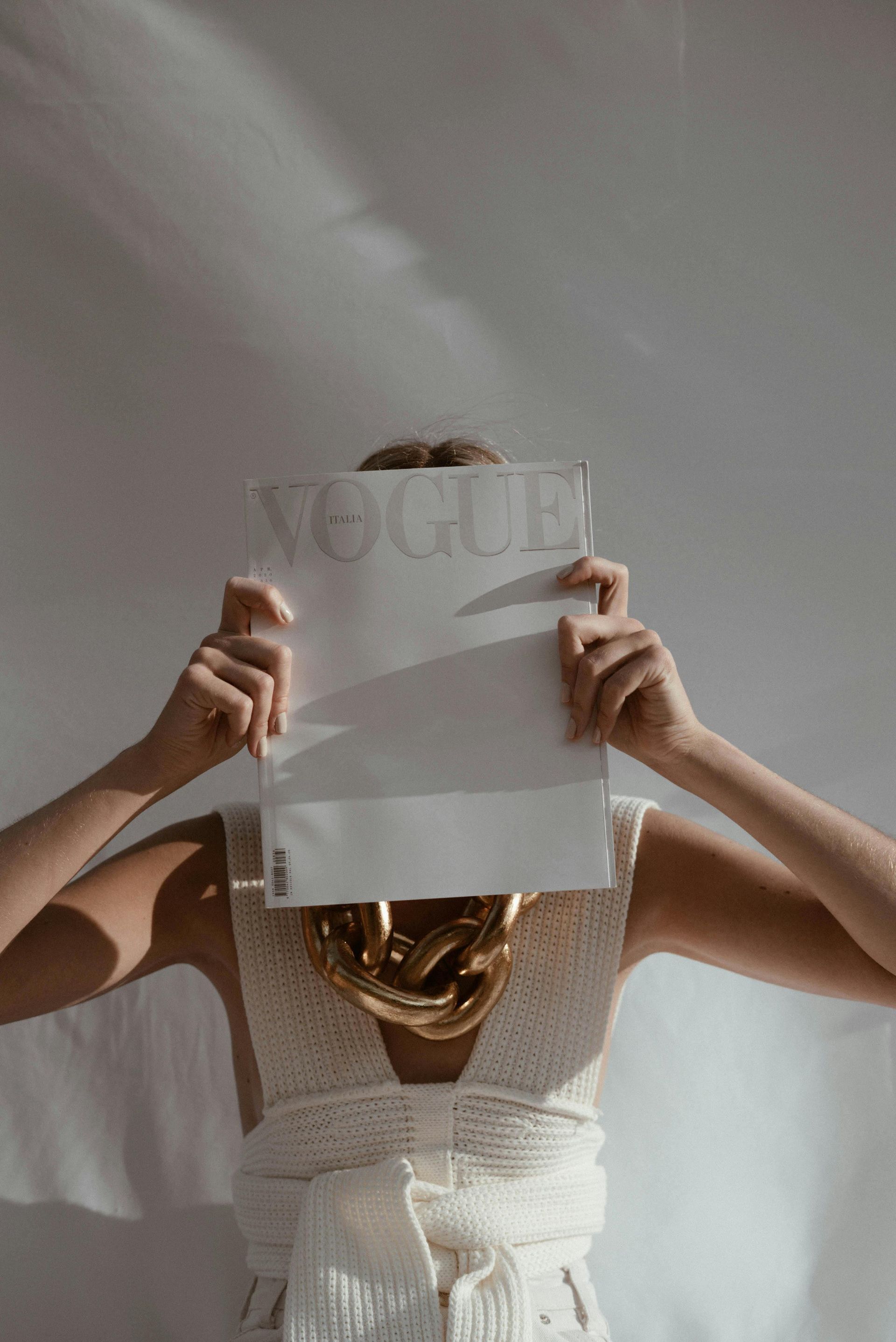 A person holding a Vogue magazine in front of their face, wearing a white crochet top and a thick, chunky gold necklace.