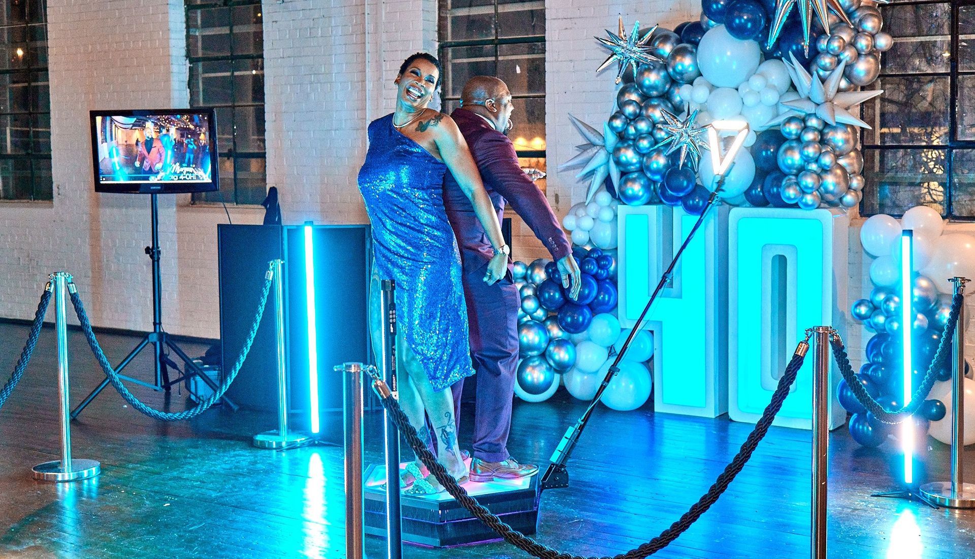 Two people pose on a platform at an event with balloon decorations and a screen.