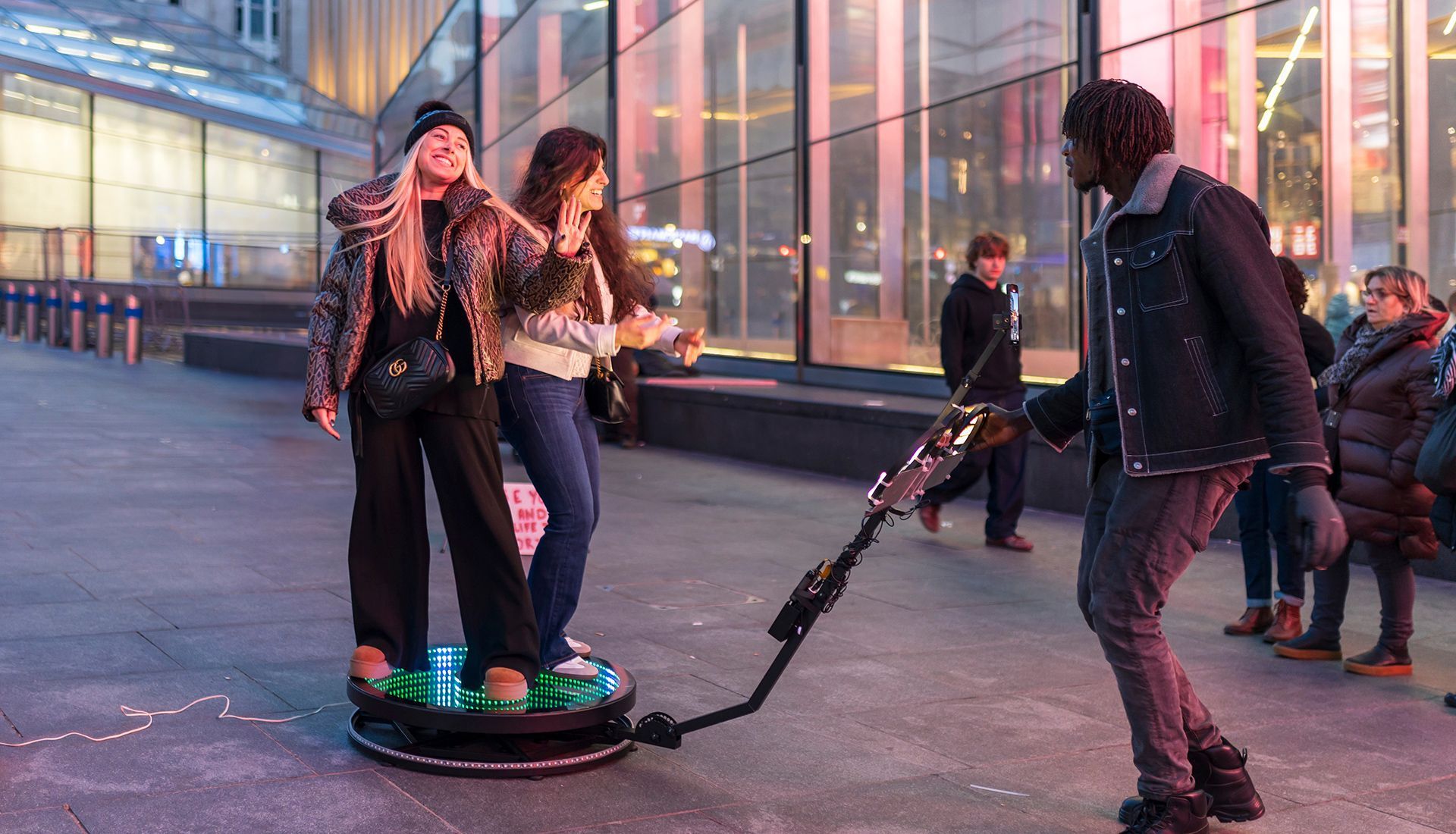 Two people stand on a lit platform as another person operates a camera apparatus outdoors.