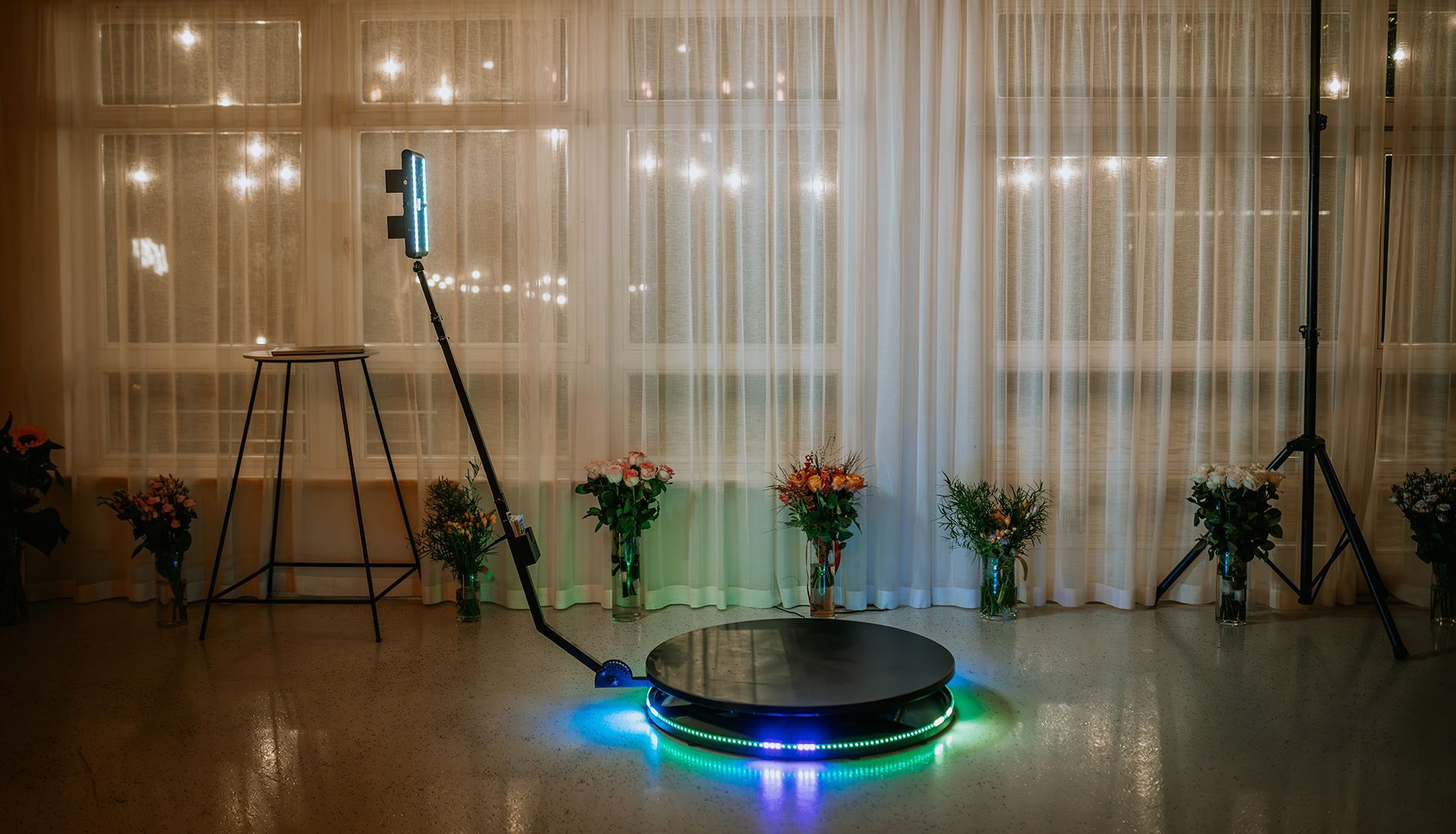 A 360-degree photo booth with a rotating arm and colorful LED base lights set up in a room with curtained windows.