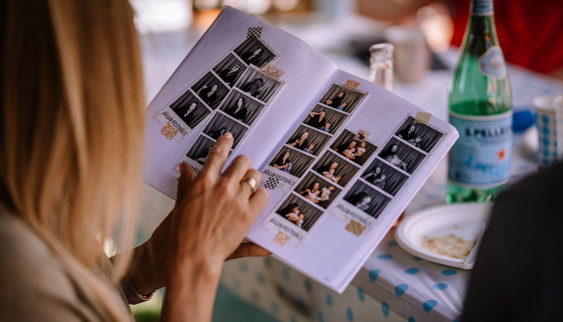 Person pointing at photos in an open photo album at an outdoor gathering, near a water bottle and plate.