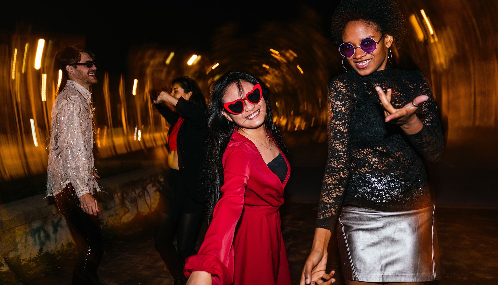 People dancing in a tunnel with blurry lights; woman in red dress smiles, another in black sparkles.